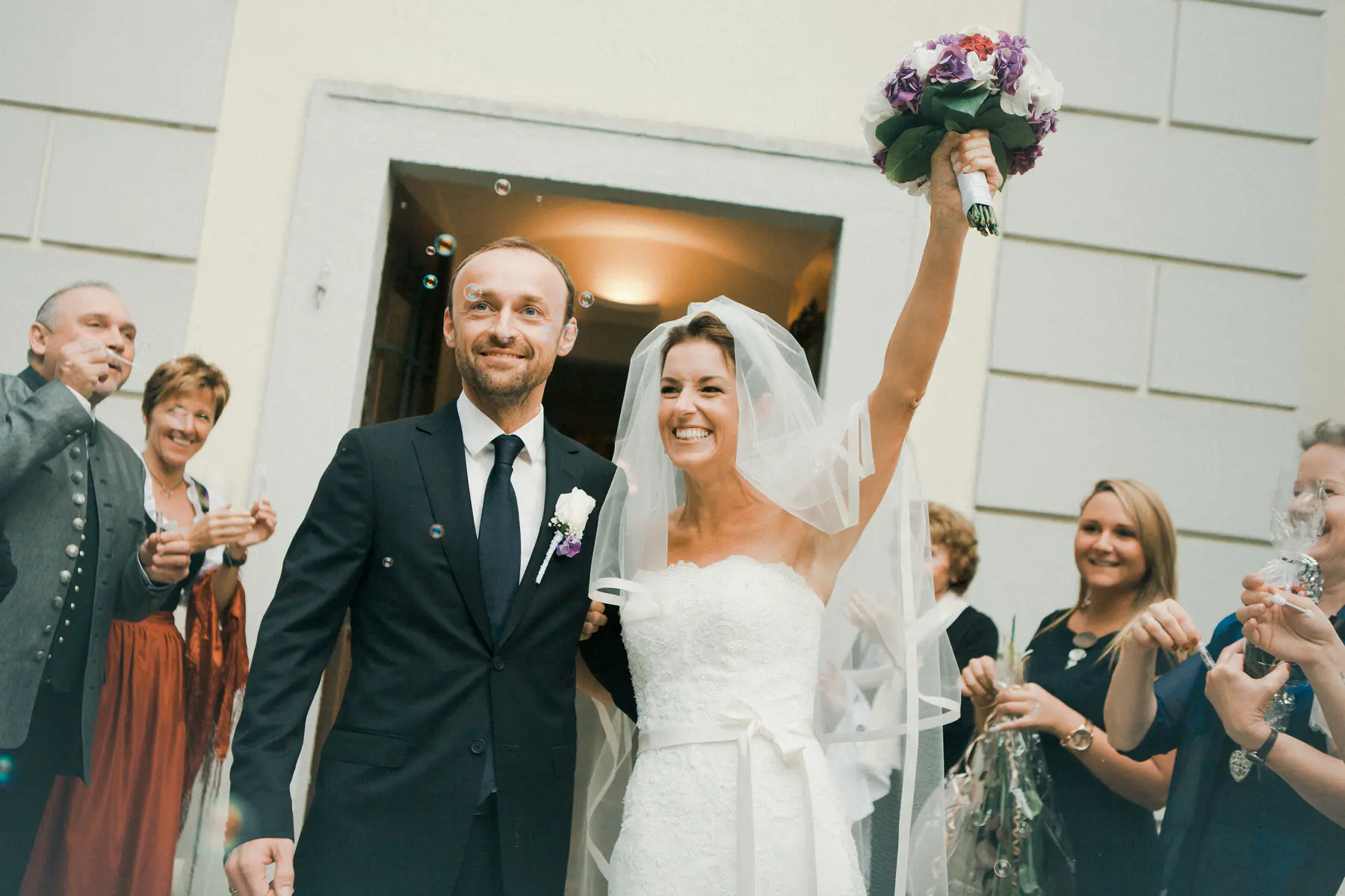 A joyful newlywed bride and groom exit a building as guests blow celebratory bubbles around them. The bride, in a white strapless gown and veil, raises a purple and white bouquet triumphantly.
