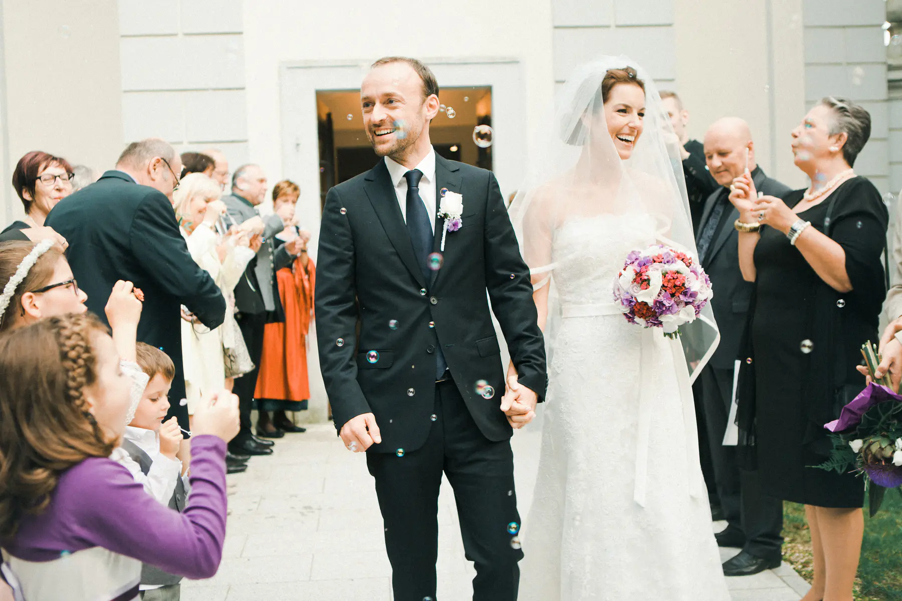A joyful bride and groom, holding hands and smiling brightly, walk out of church at Schloss Halbturn as guests line a path, blowing bubbles around them in celebration. The bride wears a white gown and veil, carrying a colorful bouquet, while the groom wears a dark suit.