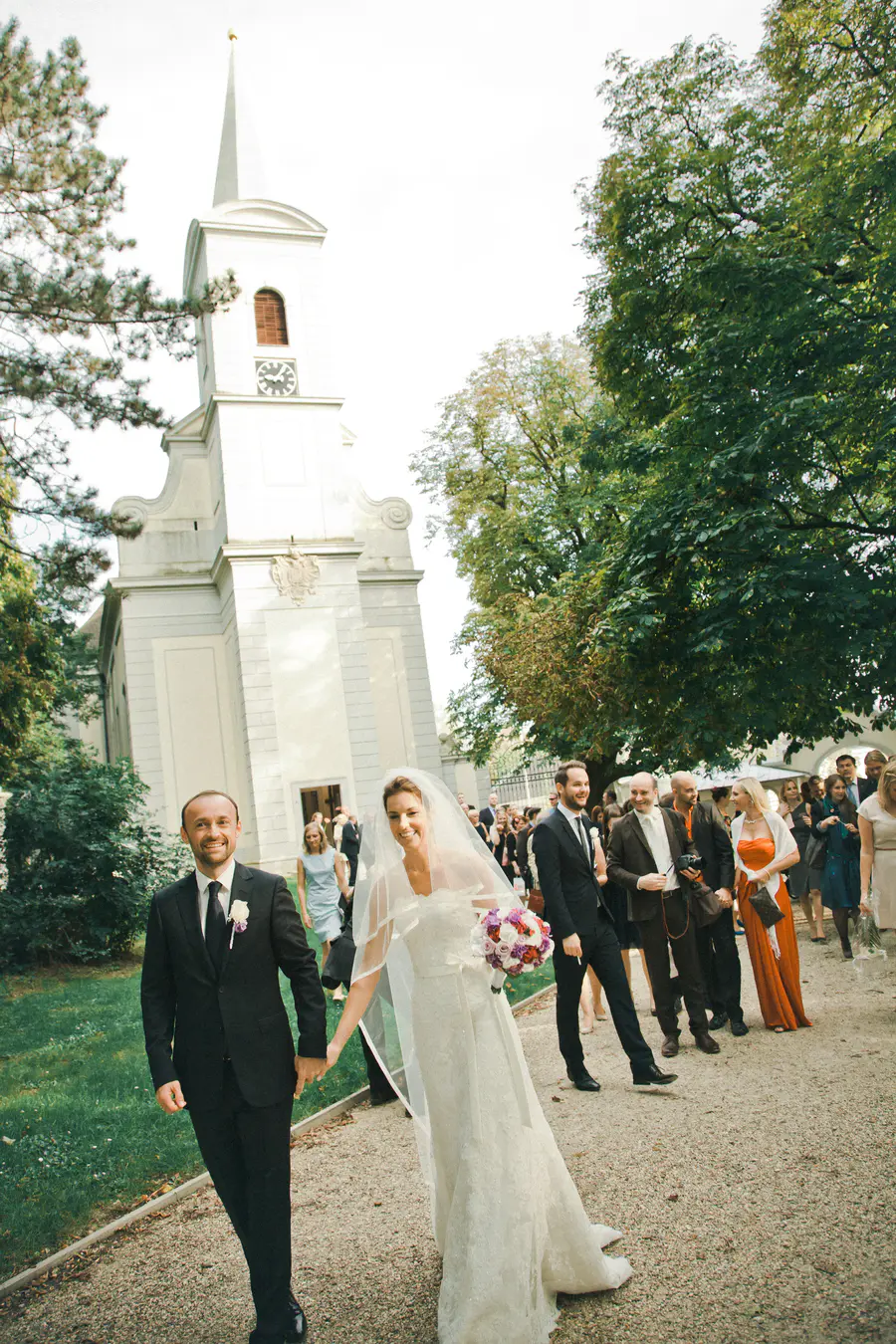 A smiling bride and groom, holding hands, walk along a gravel path after their wedding ceremony at Schloss Halbturn. The bride wears a white gown and veil and carries a purple and white bouquet, while the groom wears a dark suit. A white church with a clock tower and numerous wedding guests are visible in the background, surrounded by trees.