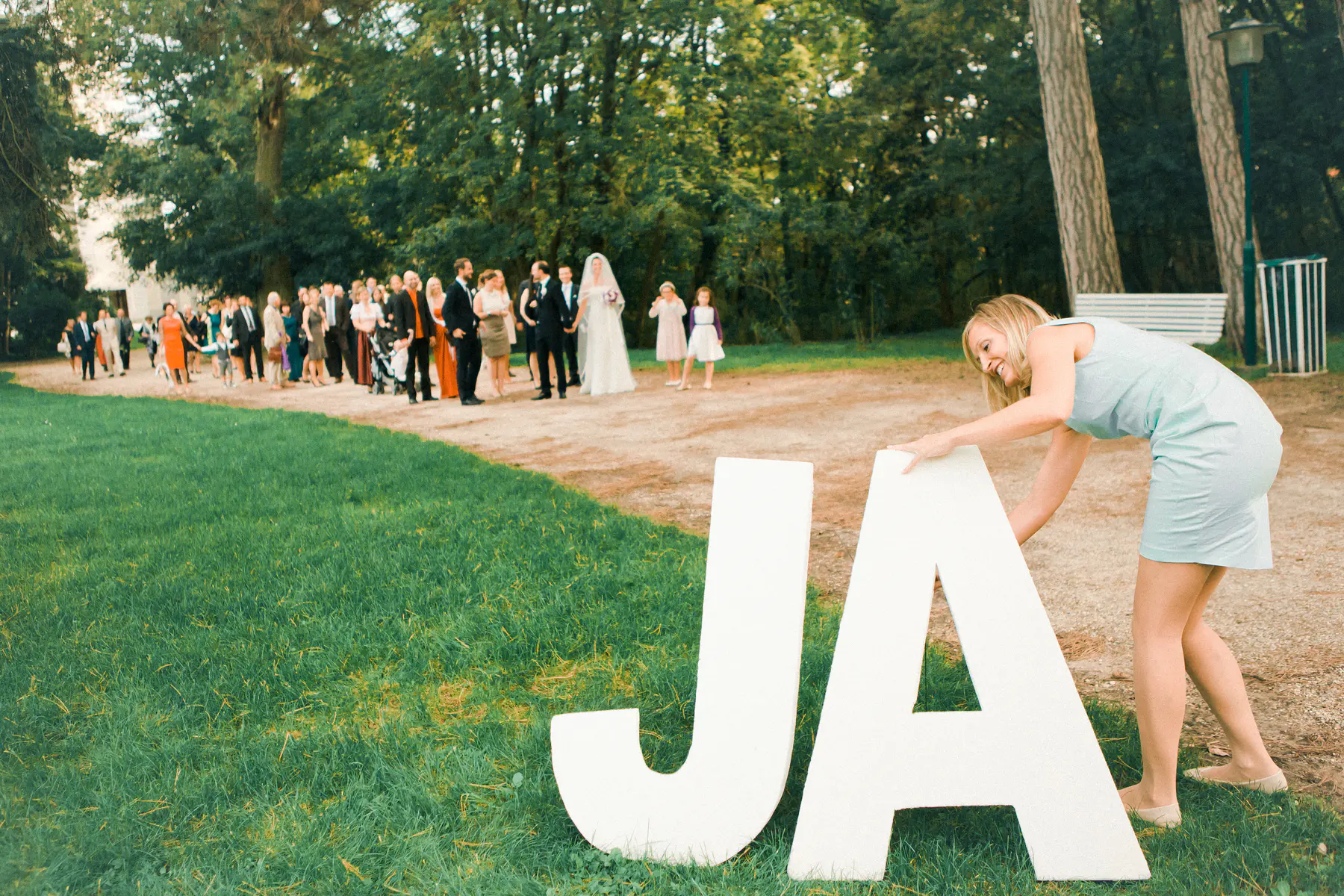 A smiling woman in a light blue dress arranges large white "JA" letters on the grass in the foreground. In the background, a wedding party including a bride and groom, along with guests, gathers on a dirt path bordered by lush green trees.