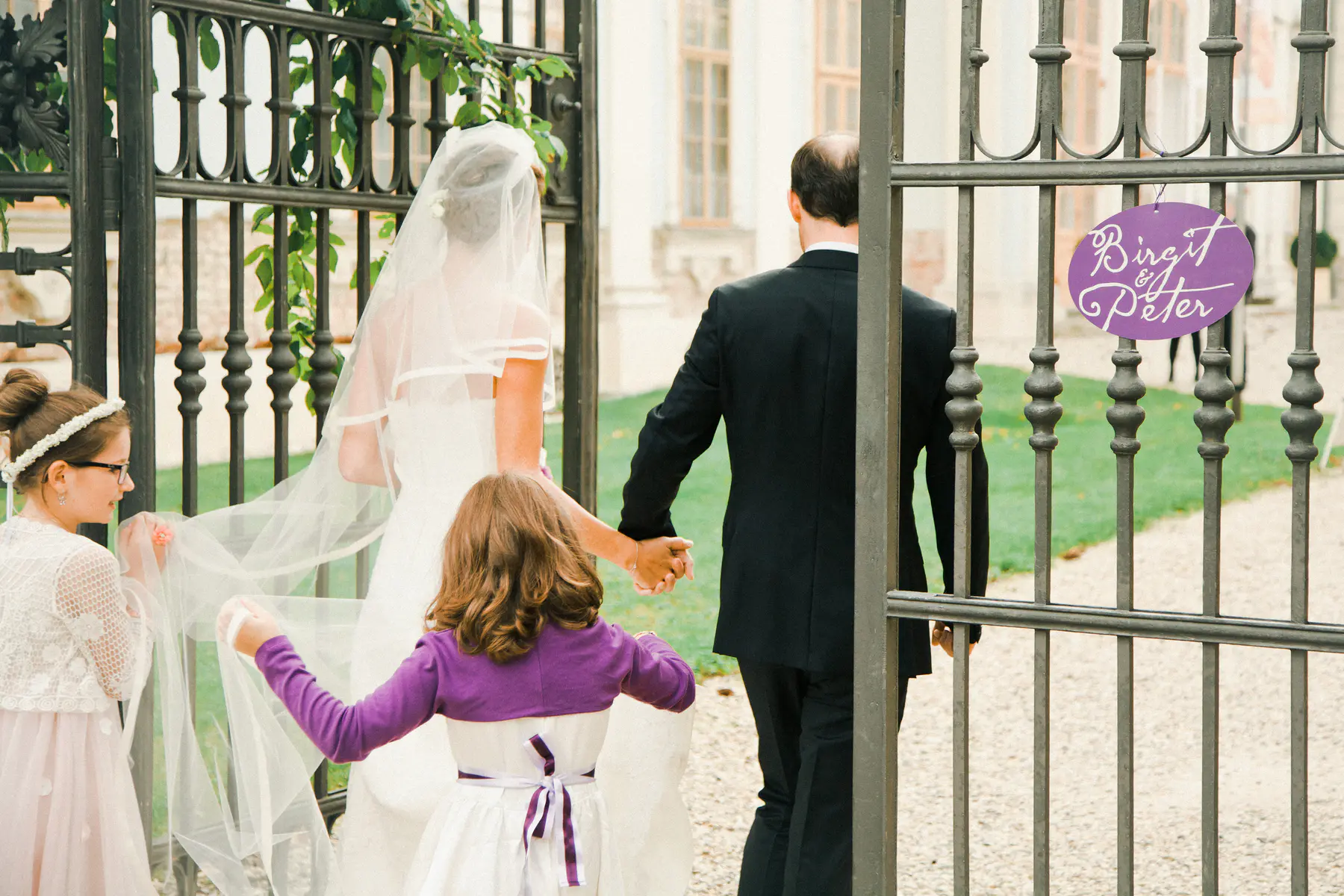 A bride and groom walk through an ornate black metal gate, viewed from behind, holding hands. A young girl in purple holds their clasped hands, while another girl in a white dress helps carry