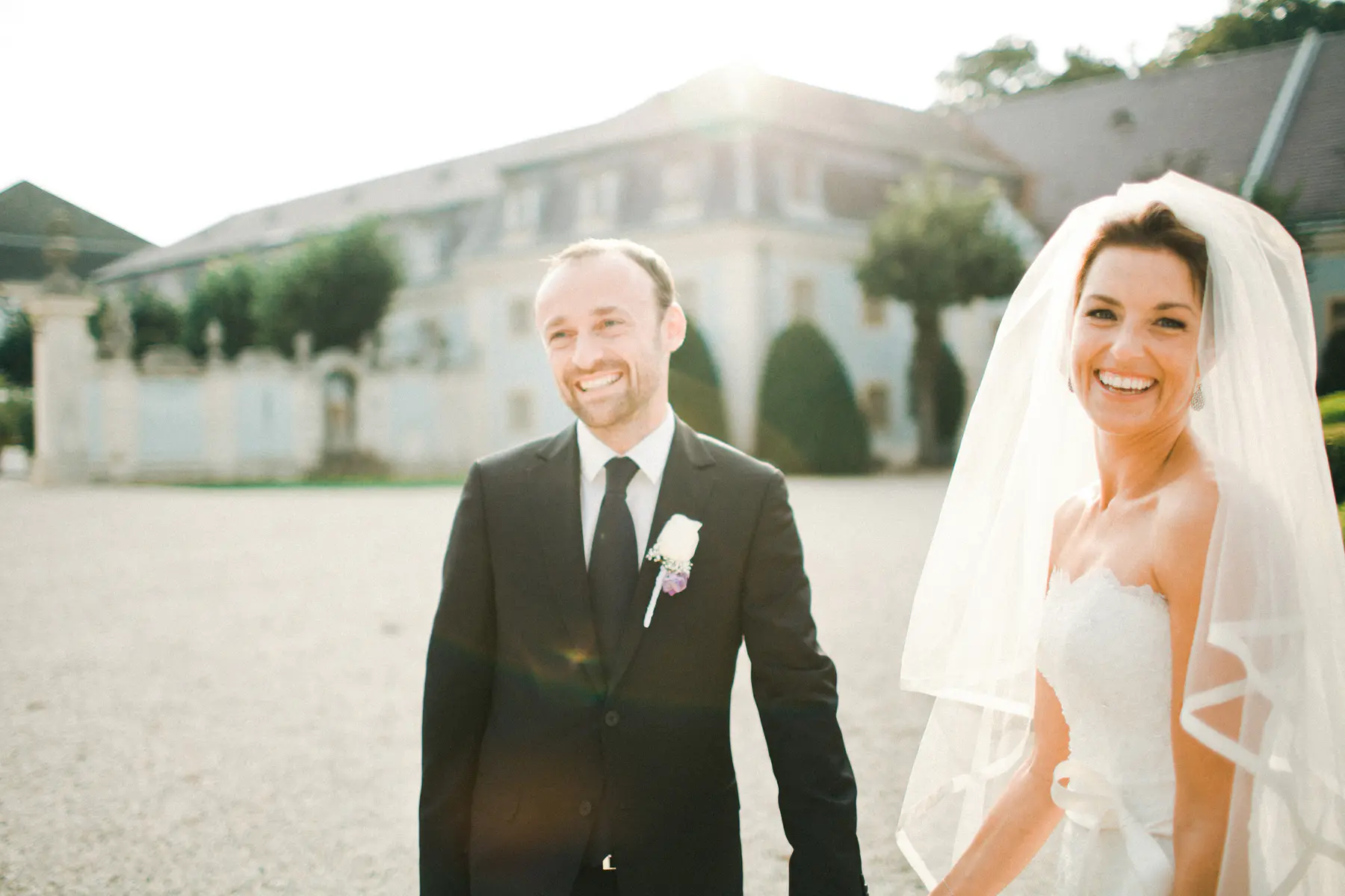 A joyful bride and groom, smiling widely, walk forward in bright sunlight with a grand estate building blurred in the background. The bride wears a white strapless gown and veil, and the groom wears