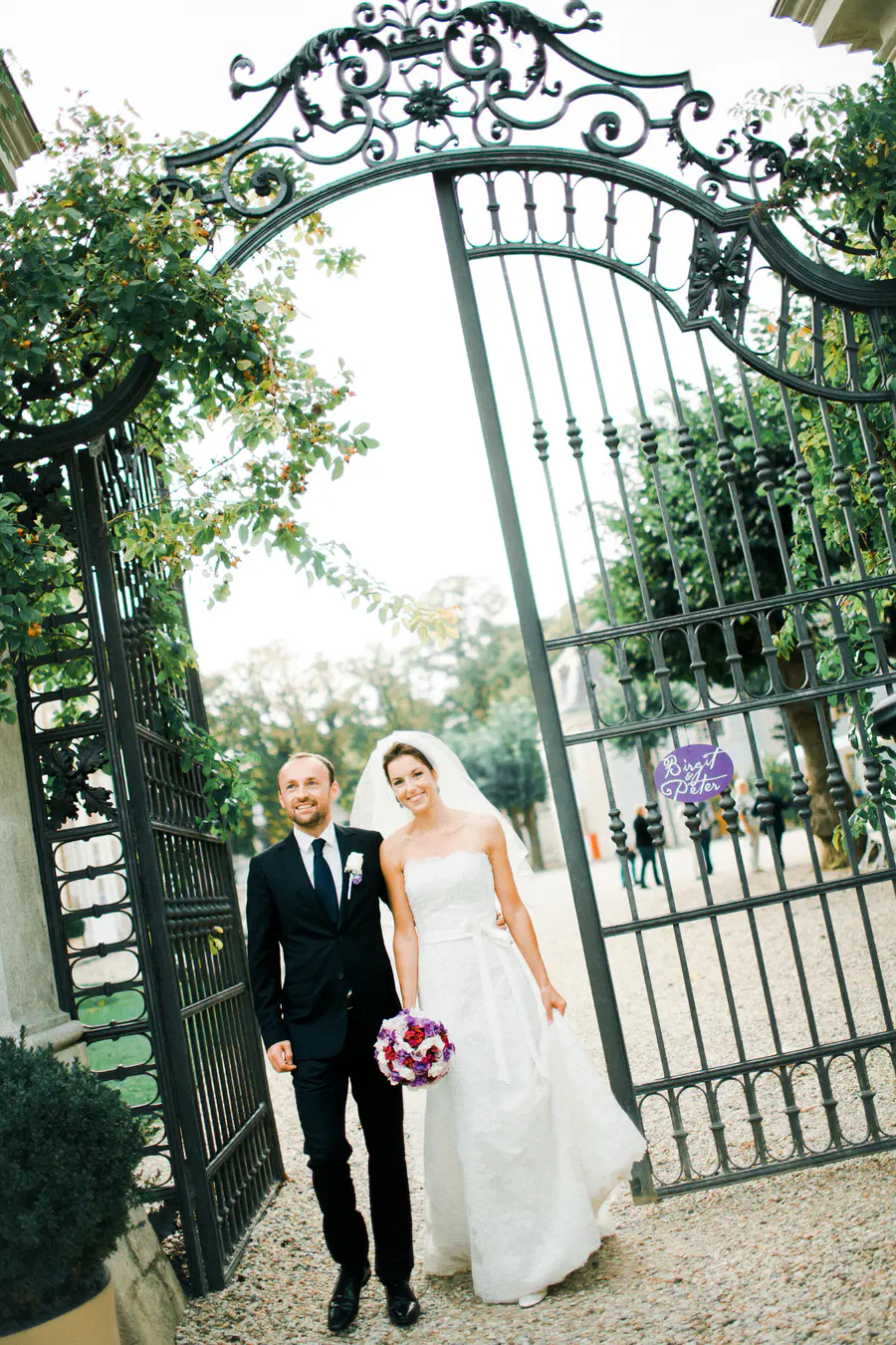 A smiling bride in a white strapless gown with a veil and purple bouquet walks alongside her groom in a black suit. They are passing through a large, ornate black wrought-iron gate adorned with green foliage. A purple sign on the gate reads "Birgit & Peter."