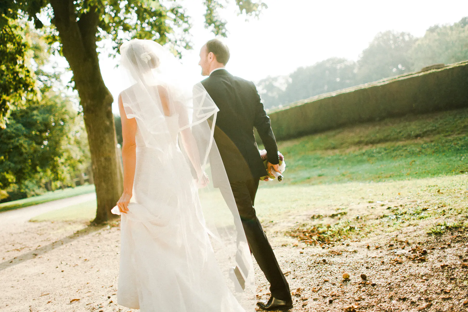 A bride in a white dress and veil walks alongside a groom in a black suit, seen from behind, as they stroll through a sunlit green park. The groom holds a floral bouquet in