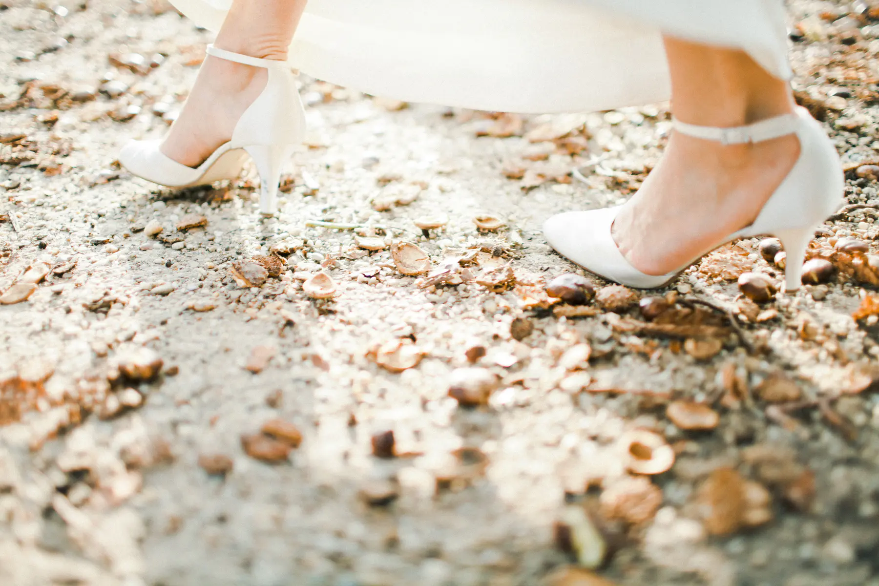 A close-up shot of a bride's feet in white heeled shoes standing on a path covered with small stones and fallen leaves.