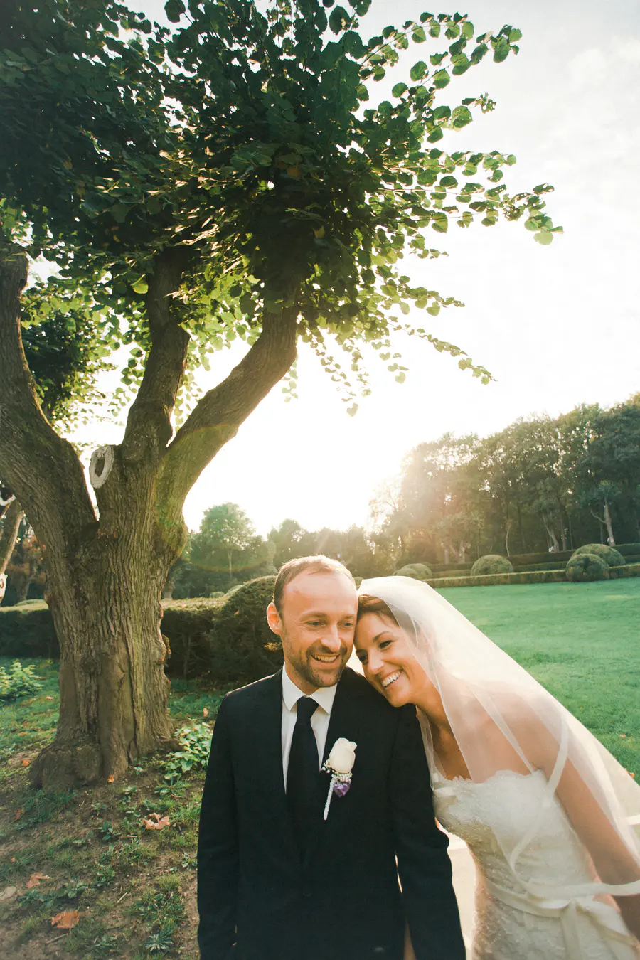 A groom and bride smile at each other under a tree on their wedding day.