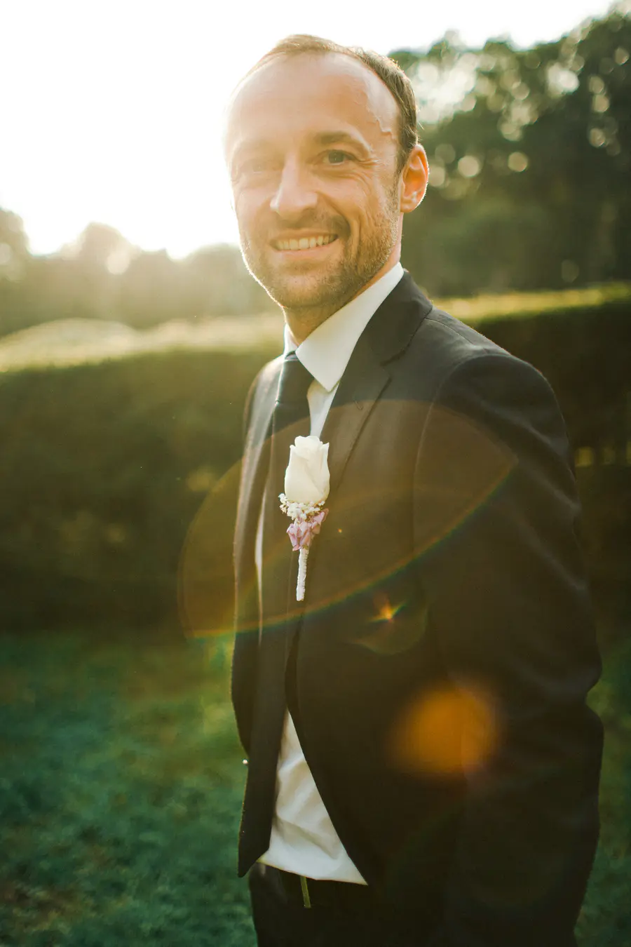 A man in a black suit and tie smiles at the camera, with a white rose boutonnière pinned to his lapel. He stands outdoors with soft sunlight and trees in the background.
