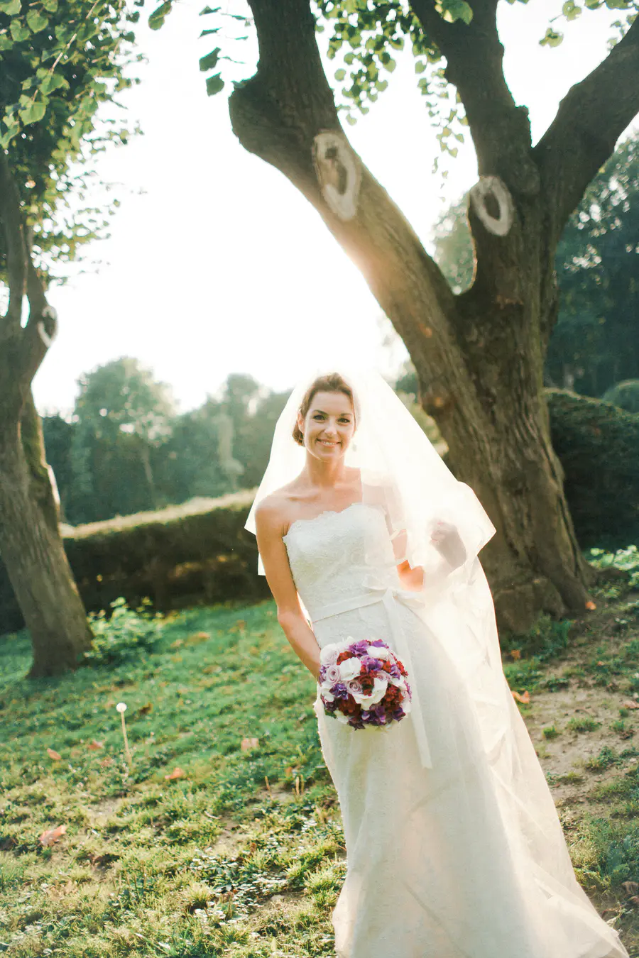 A bride in a white lace wedding dress and veil smiles at the camera, holding a bouquet of red, purple, and white flowers. She is standing in a grassy area with trees in the background.