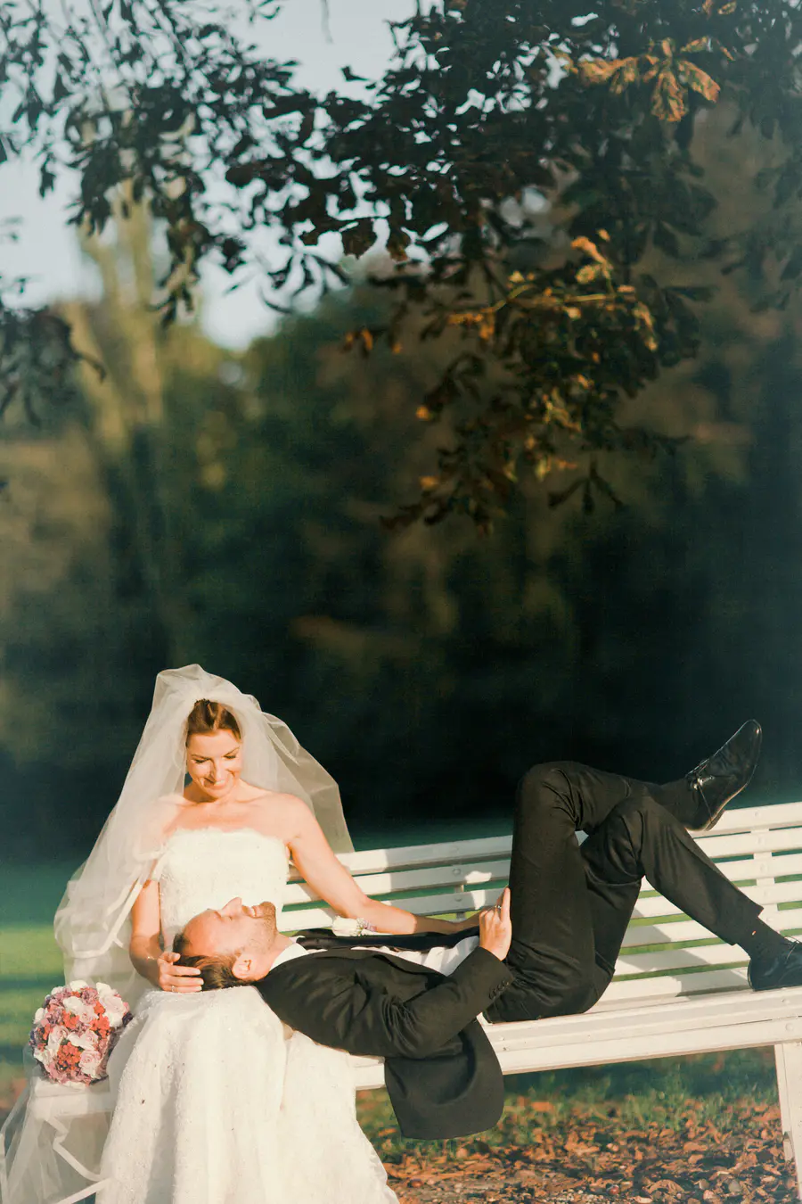 A bride in a white wedding dress and veil rests her head on the lap of her groom, who is lying on his back on a park bench.