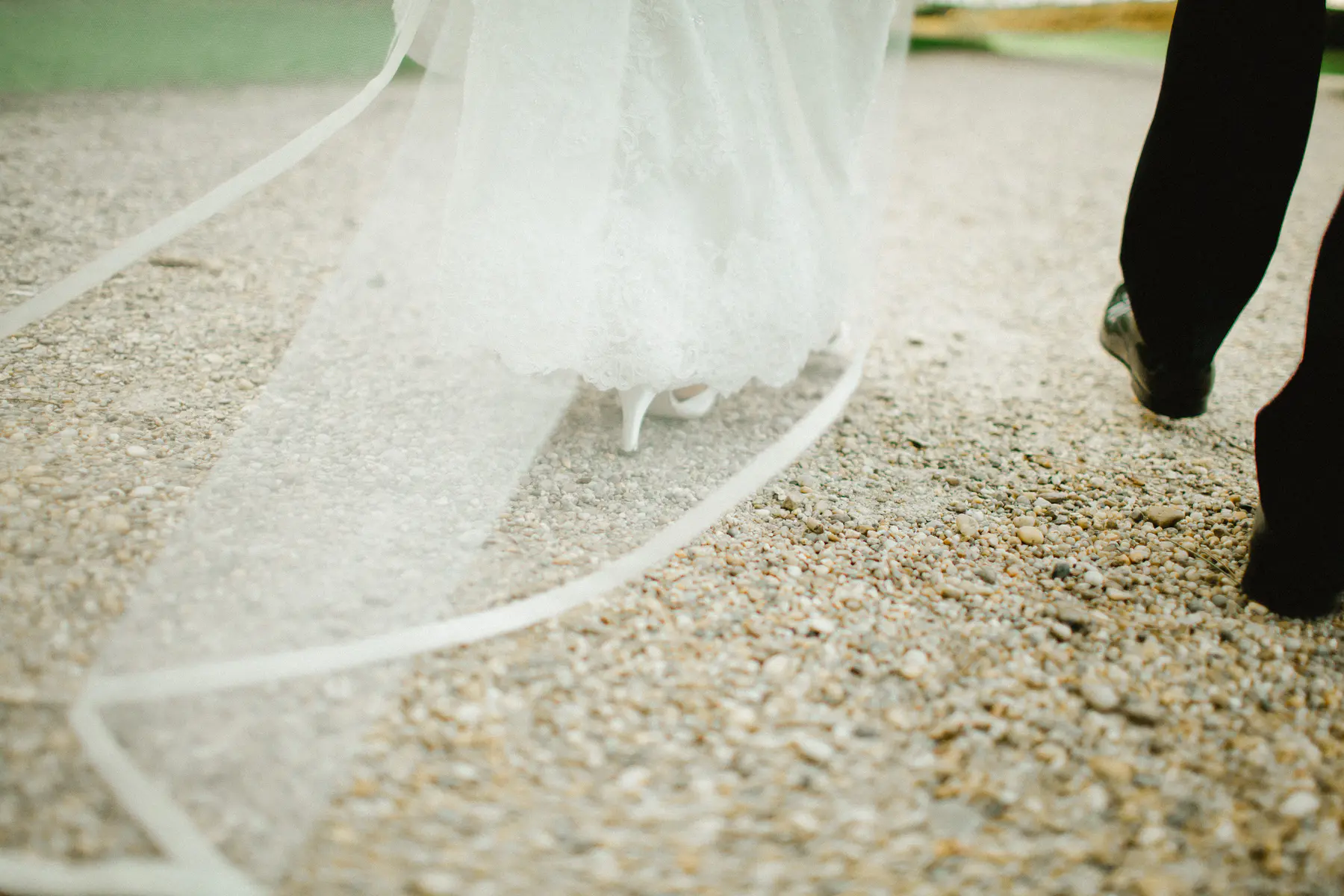 A bride in a white lace dress and white heels walks on a gravel path, her veil trailing behind her, next to a person in black pants and dress shoes.
