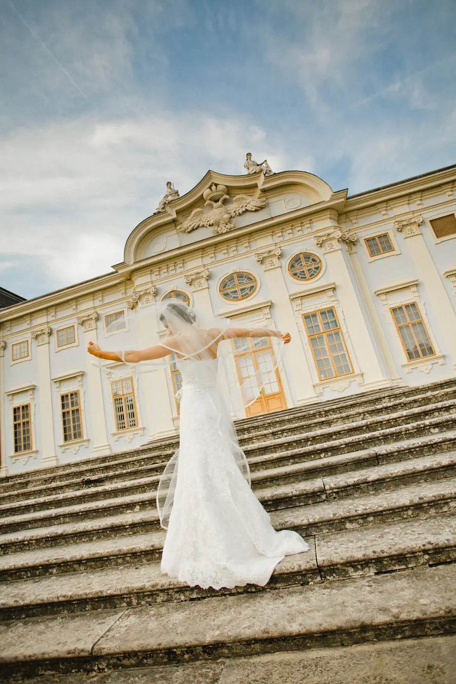 A bride in a white lace wedding dress and veil stands on a wide, stone staircase, with her arms outstretched to the sides. Behind her, a grand, light-colored building with multiple windows and decorative architectural details looms against a cloudy sky.