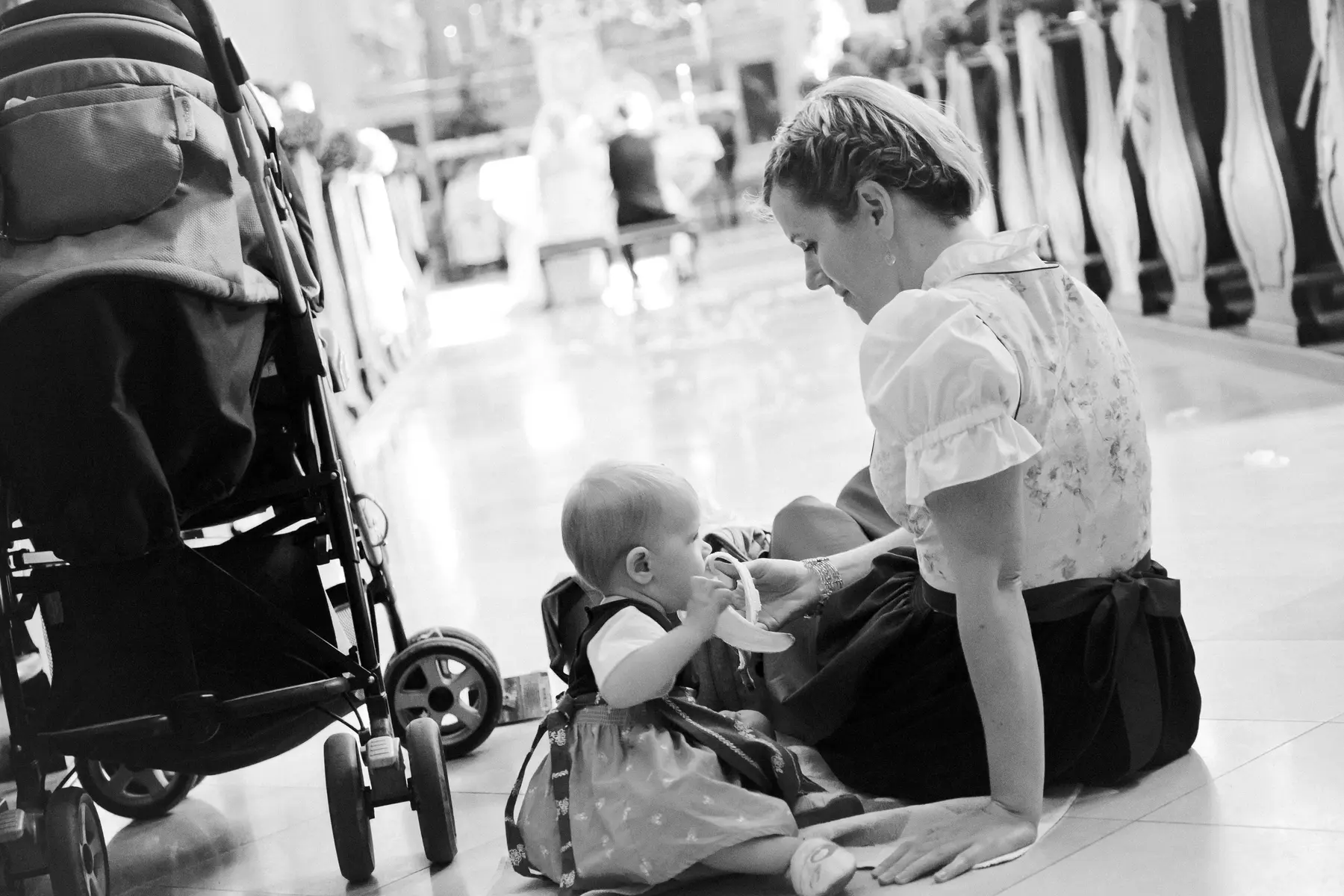 A black and white photo of an adult woman with braided hair, wearing a traditional Dirndl, sitting on a tiled floor and smiling gently at a baby in a matching outfit. The baby holds