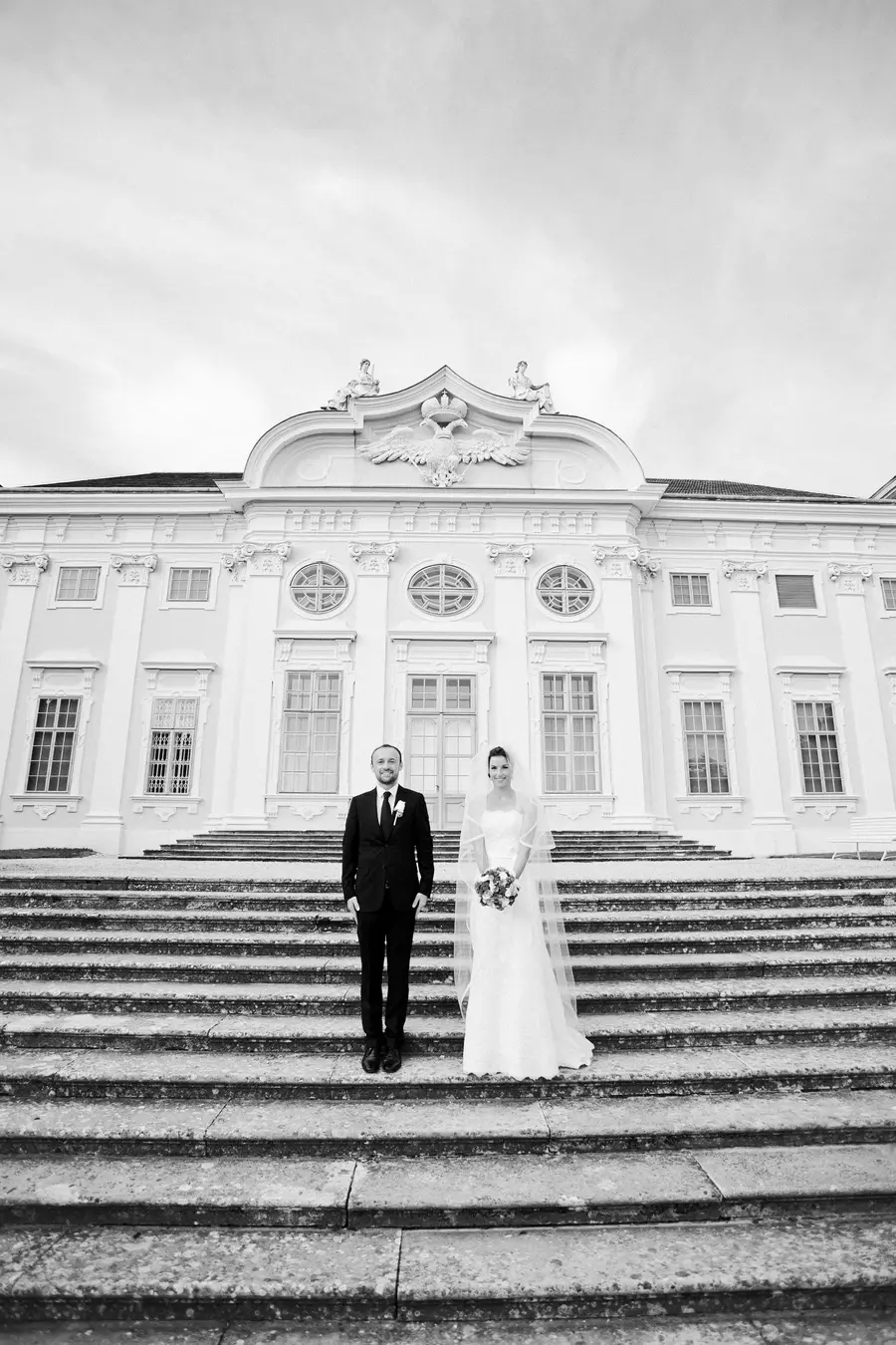 A black and white, wide shot captures a newlywed couple standing on the steps of a grand, ornate building. The groom, on the left, wears a traditional black suit and tie, while the bride, on the right, is in a white wedding gown with a veil and holds a bouquet. They are both looking at the camera.